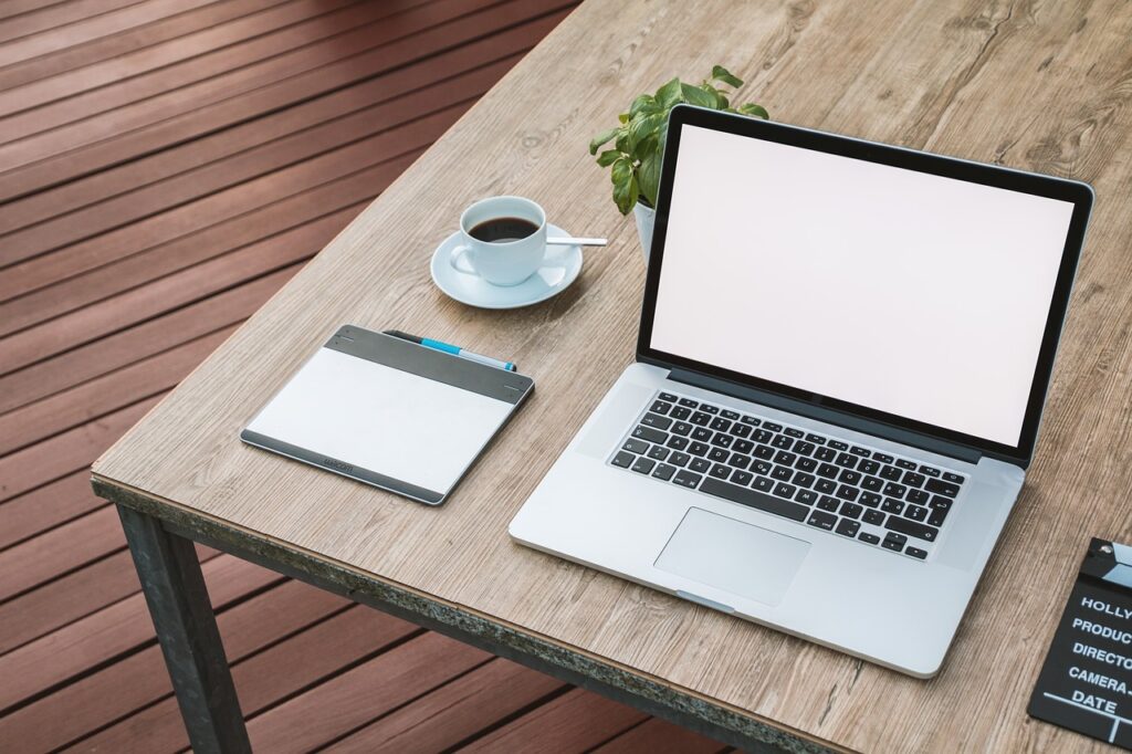 Auf einem Holztisch stehen ein Laptop, eine Kaffeetasse, ein Grafiktablet und eine kleine grüne Pflanze